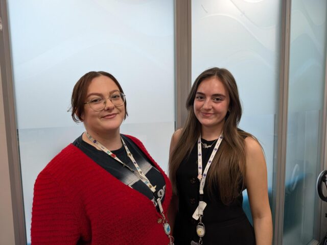 two people stand next to each other against a frosted window. One has brown hair tied up and has glasses on. The other has long brown hair, and is wearing a black sleeveless shirt