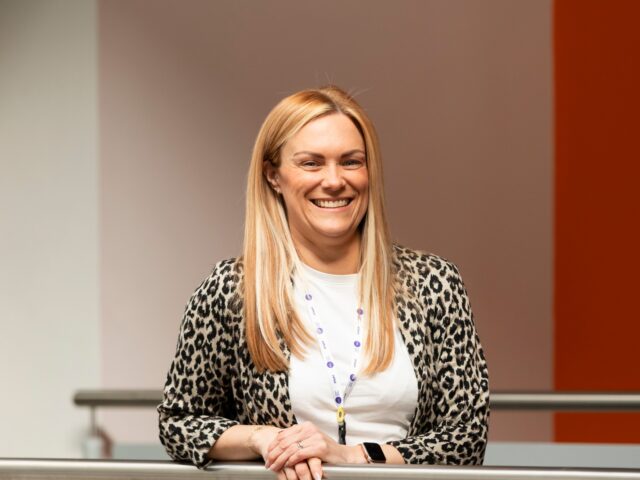 a person with chest-length blond hair smiles at the camera. They have a leopard print suit jacket on