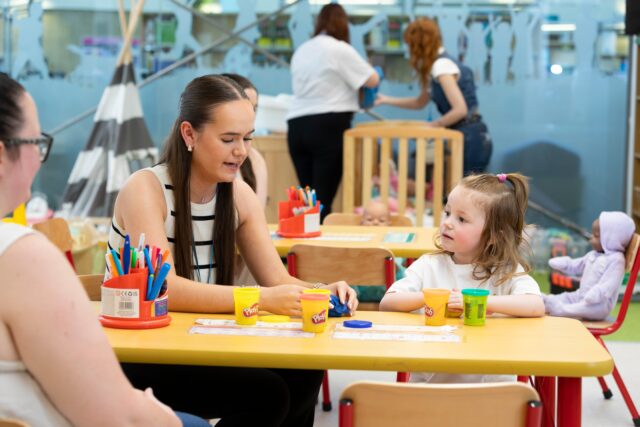 a person sits in a classroom setting, looking at play dough on a table with a little girl.