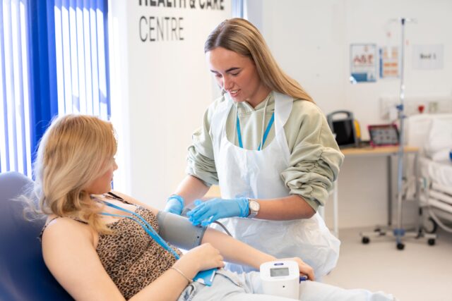 a person takes another persons blood pressure in a hospital setting. Both people have long blond hair and one has medical gloves on and a medical apron