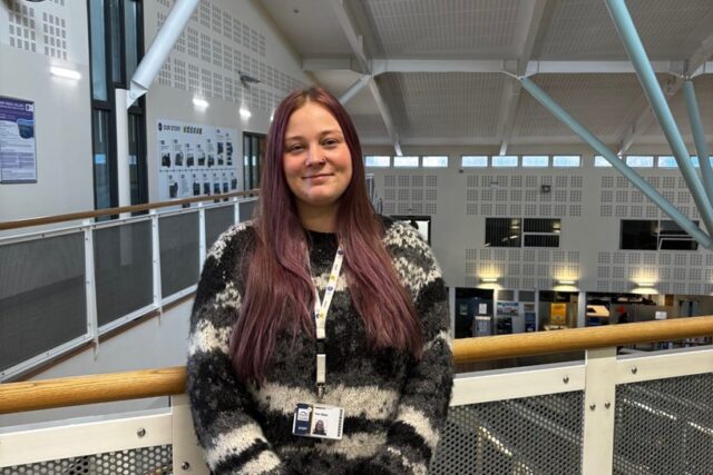 a person with long purple hair smiles at the camera. She appears to be on the balcony of a bigger building, and has a staff lanyard on, and a black and white striped jumper