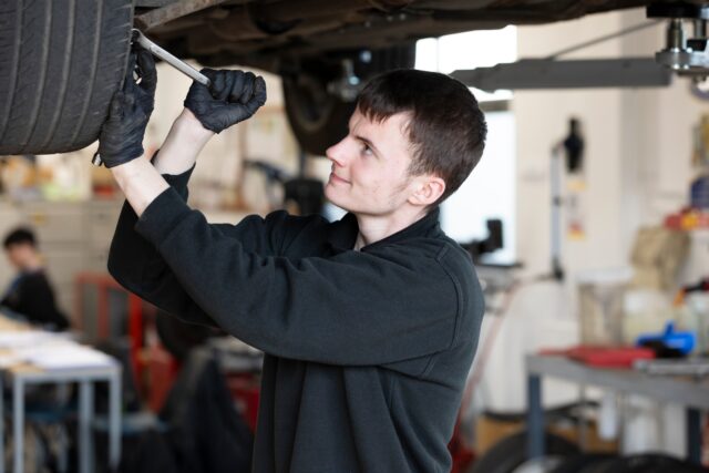 a person with short dark hair and dark overalls holds a tool and is underneath a car in a workshop setting