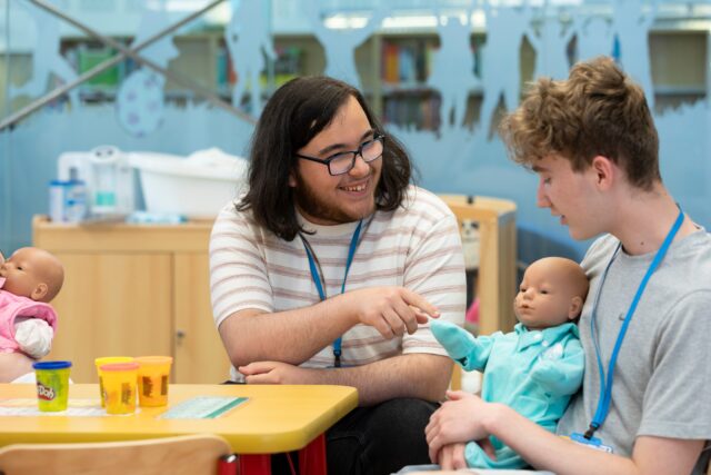 two people sit at a table, one is holding a pretend doll. One person has a striped t shirt on, long brown hair and dark rimmed glasses, and they are smiling at another person who has short blonde hair
