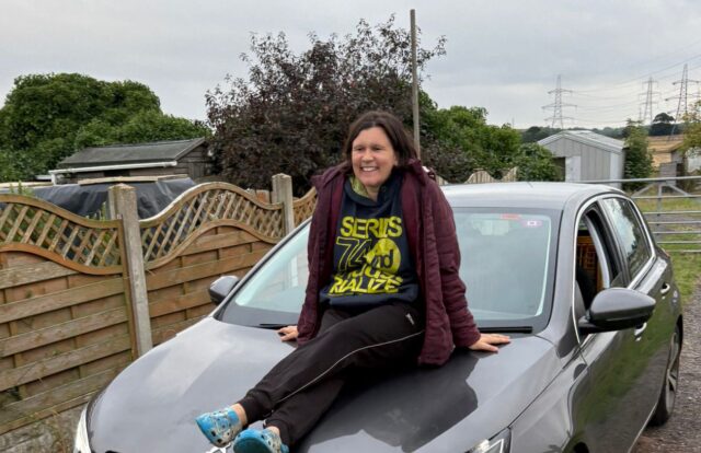 someone sits on the bonnet of a car, smiling at the camera
