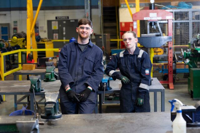 two people in industrial workwear stand smiling at the camera against NETA Training background