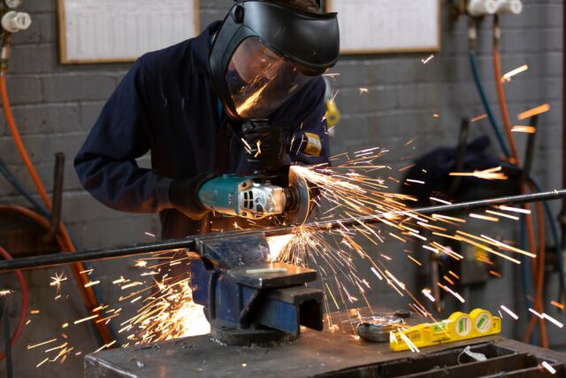 someone in industrial workwear wears a large protective helmet and mask and cuts pipe. There are sparks flying