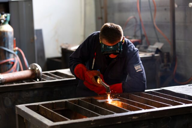 someone in protective industrial workwear is manufacturing / fabricating. Their face is not visible but you can see the heat and sparks from the tool they're using