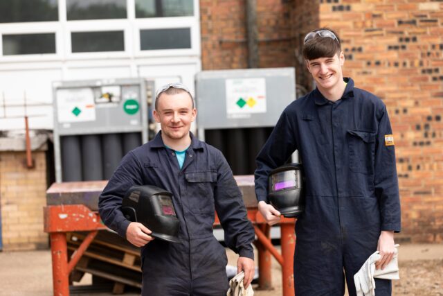 two people stand looking at the camera against a brick building. They both have short dark hair and are dressed in industrial workwear. One is holding a protective helmet under his arm.