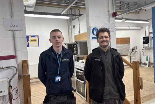 two people stand in a learning space designed for plumbers. Both people have short dark hair and are wearing dark clothes suited to the plumbing trade.