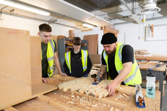 3 people stand over pieces of wood and they're using a tool on it. Two of the people are students and one a tutor. The tutor has a beard and a beanie hat on. One student has short hair and the other has long hair in a ponytail and dark rimmed glasses. All 3 are wearing high vis