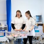 two people dressed in white look over a transparent hospital baby bed. There is a baby mannequin in the bed, and they look over it in what looks like a hospital setting