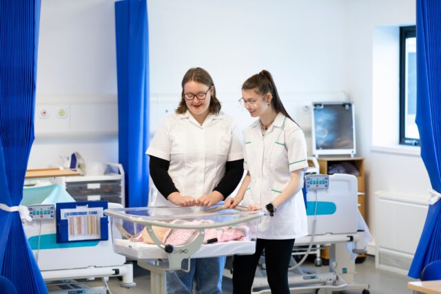 two people dressed in white look over a transparent hospital baby bed. There is a baby mannequin in the bed, and they look over it in what looks like a hospital setting