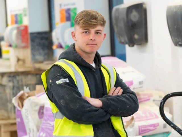 a person with short blonde hair looks at the camera. Their arms are crossed, and they're wearing a black hooded sweatshirt and a short sleeved high vis jacket