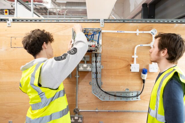 two people stand against a wooden wall with and electrical circuit set up. The two people's faces are out of view but they are working on the circuit, both wearing short high vis vests