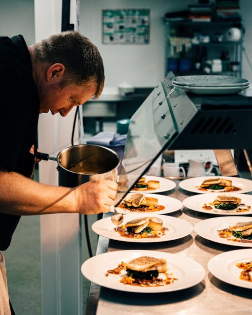 a person with short dark hair stands at a chefs counter, pouring sauce onto dishes with food in
