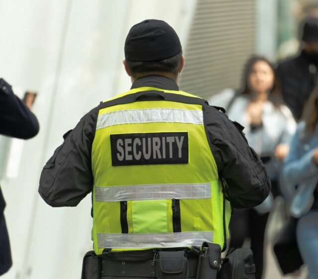 the back of someone's head and torso. They have a high vis jacket on with SECURITY in large letters