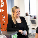 a person smiles at something off camera and has a long blond ponytail. They are dressed in black - a top and a blazer.