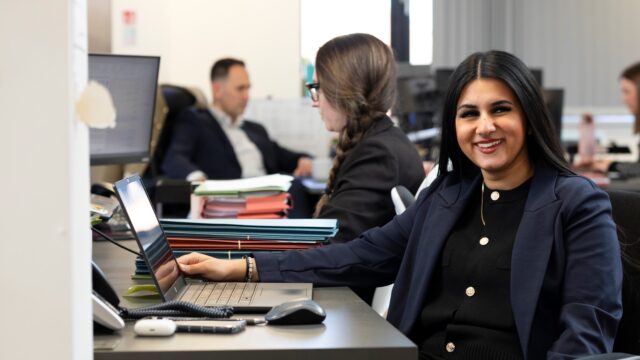 a person in a navy suit smiles at the camera. they are sat at a desk and have long dark hair