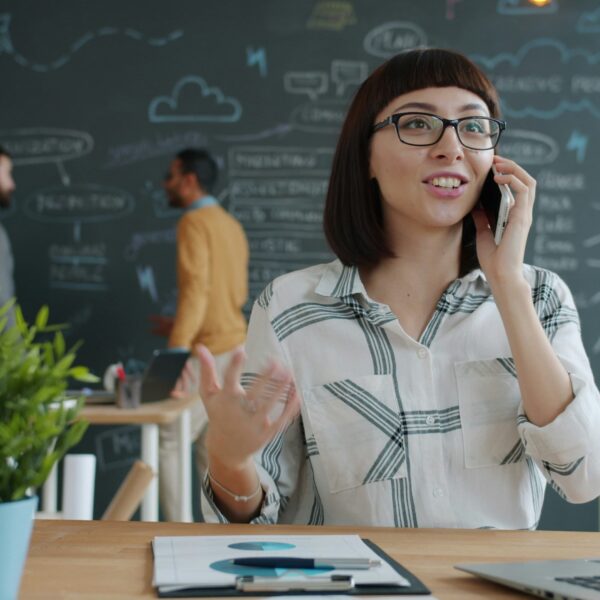 a person sits at a table on the phone smiling. They have a laptop infront of them and appear to be in a cafe
